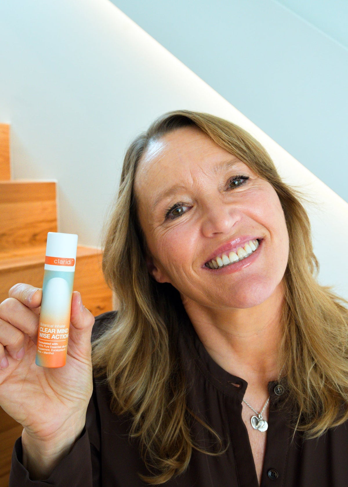 A smiling woman with blonde hair holds up a small cylindrical tube with a white cap and orange/white label, standing in front of a staircase and light blue wall.